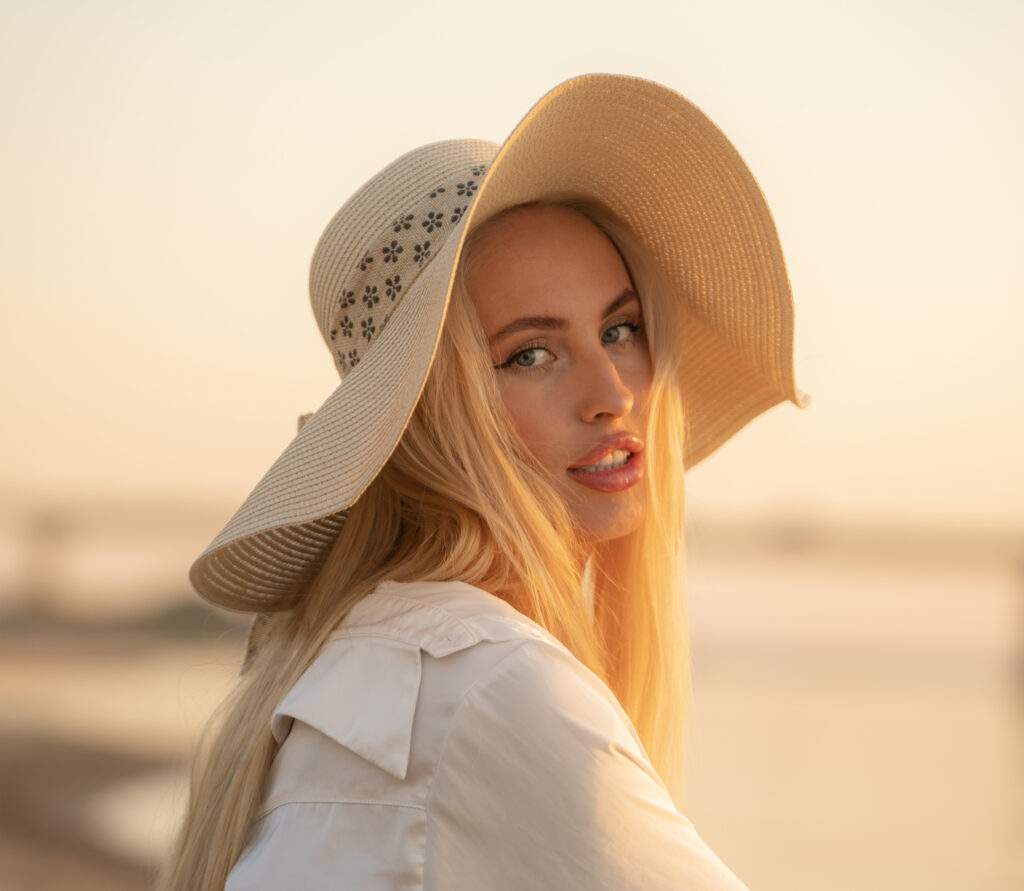 A woman with a large hat relaxes on the sandy river bank as the sun sets, creating a serene atmosphere with reflections in the water.
